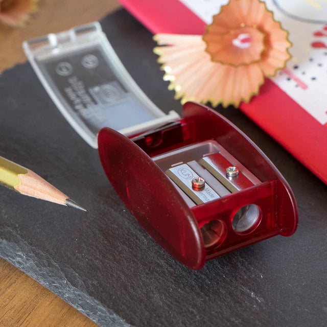A translucent red pencil sharpener with two metal sharpening blades, shown open on a dark slate surface with a pencil, notebook, and wooden shavings in the background.