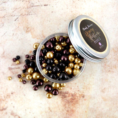 An open round tin filled with glossy pearl beads in metallic gold, deep purple, and black, with some beads spilled onto a light stone surface. The silver lid has a round black label reading 'Breathe Jewelry Pearl Beads'.