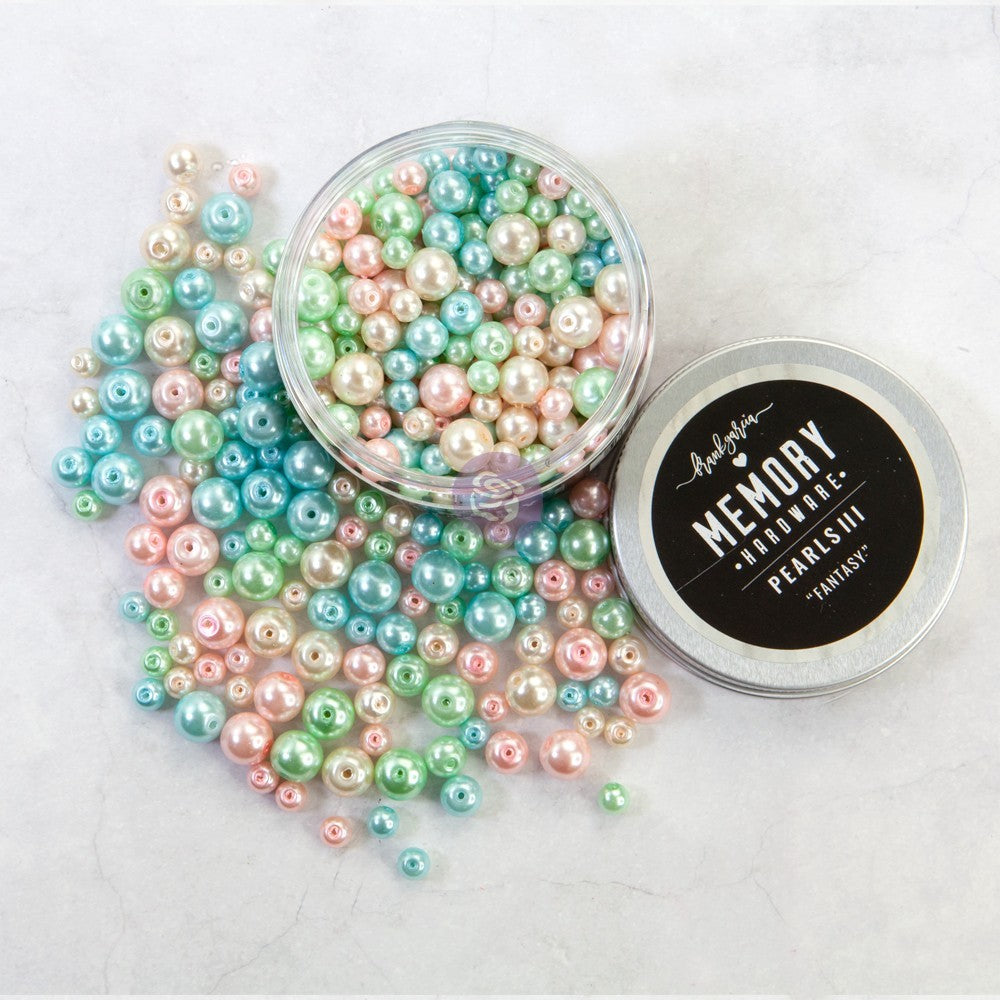 A clear round container filled with iridescent pearl-like beads, with several beads spilled around it on a light marble surface. A silver lid with a black label reading 'Memory Beads Pearls III Fantasy' is placed beside the container.