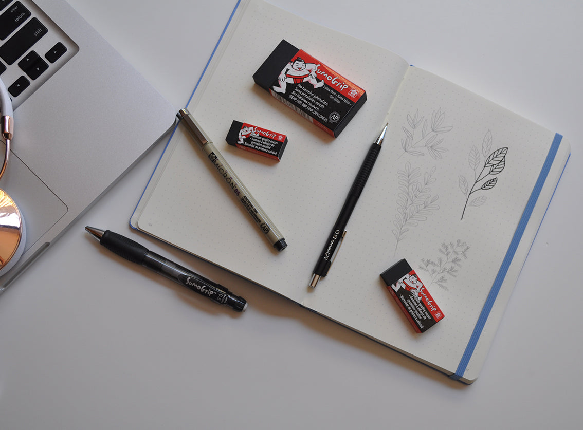 An open sketchbook or notebook with blue binding, placed on a white desk alongside pens, an eraser, and pencil. Part of a laptop is visible at the left edge.